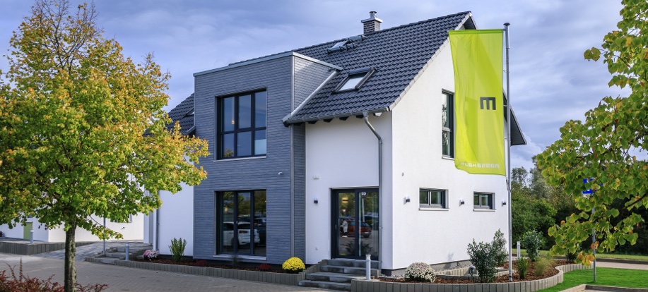 Modernes Einfamilienhaus mit großen Fensterflächen, grau-weißer Fassade und grünem Banner vor blauem Himmel.